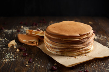 stack of pancakes, folded pancake, sprinkled with berries, cookies, sugar powder and caramel syrup on parchment on a dark wooden table