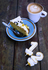 Slice of birthday cake witn candle numbers one and two with cup of latte on wooden table. Text space