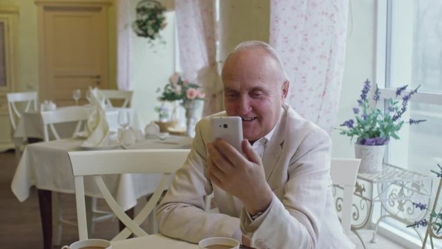 Pan of cheerful senior man sitting in cozy cafe and chatting with someone over video call on mobile phone
