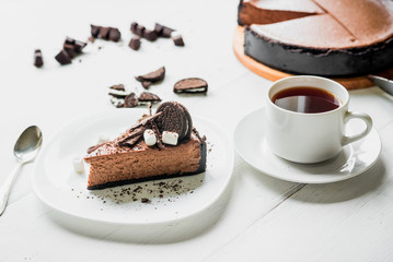 Chocolate cheesecake with pieces of chocolate, cookies and marshmallow on a white plate next to a cup of tea on the table
