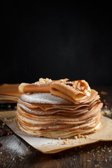 two stacks of pancakes, two folded pancakes, sprinkled with berries, cookies, sugar powder and caramel syrup on parchment on a dark wooden table