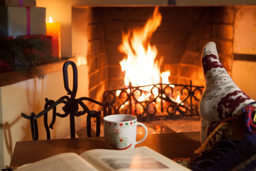 A woman in warm knitted socks with a cup of hot mulled wine and an old book in front of the...
