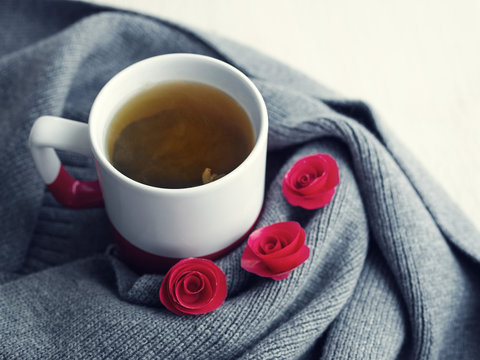 Cup of hot tea and decorative red flowers on a scarf on a white wooden surface of a table. Romantic background