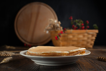 a pile of pancakes, spikelets, a round cutting board, a basket with branches with red berries and berries on a dark wooden table
