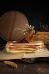 a pile of pancakes, spikelets, a round cutting board, a basket with branches with red berries and berries on a dark wooden table