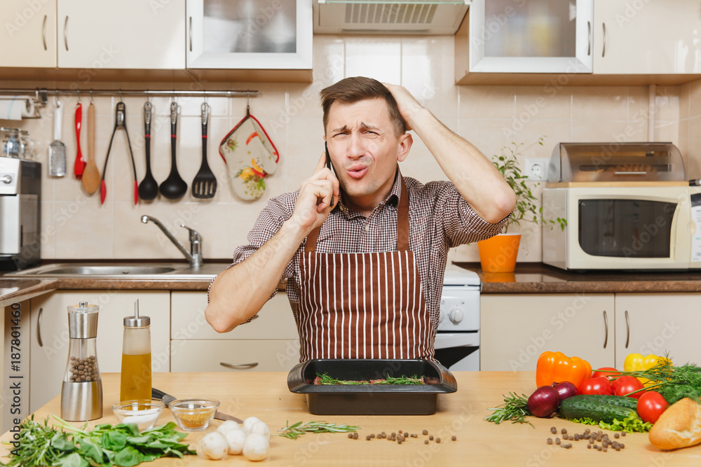 Wall mural Perturbed stress young man in apron sitting at table with vegetables, talking on mobile phone, cooking at home preparing meat stake from pork, beef or lamb, in light kitchen with wooden surface.