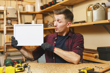 Handsome caucasian young man in plaid shirt, gloves working in carpentry workshop at wooden table place with blank frame, different tools. With empty place for text. Copy space for advertisement.