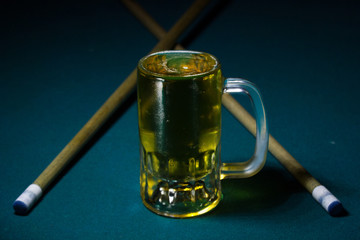 cold beer mug on a green billiard table with two cue stick in V shape 