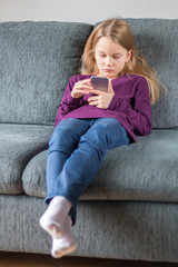 Mädchen tippt auf einem Smartphone und sitzt auf einem grauen Sofa