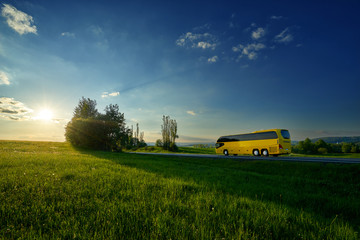 Yellow bus traveling on the road between green meadows in a rural landscape at sunset