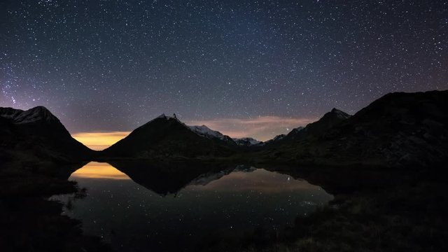 Starry sky and Orion Time Lapse on the Alps, beyond snowcapped mountain ridge, reflection on idyllic alpine lake.
