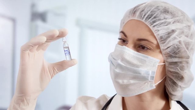 Professional female doctor in mask, cap and gloves standing in hospital room holding ampoule with new drug treatment. Woman physician at work. Laboratory employee making scientific research