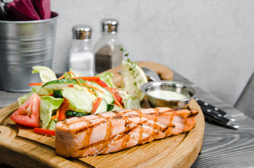 Grilled salmon steak with tartar sauce and fresh vegetables served on a wooden table at restaurant. Grey background with space for text. Pepper and salt, Knife and fork, metal