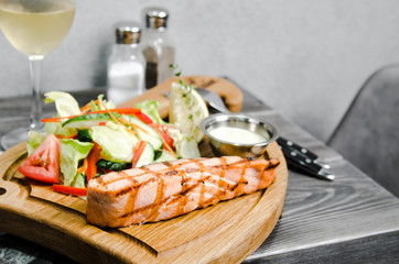 Grilled salmon steak with tartar sauce and fresh vegetables served on a wooden table at restaurant. Grey background with space for text. Pepper and salt, Knife and fork, metal