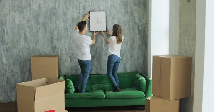 Young happy millennial couple students move into their first new owners home. Simple background of urban apartment, they frame and decide on decoration on walls, painting and map
