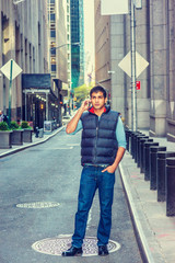 Modern City Life. East Indian American Man wearing long sleeve T shirt, blue down vest, jeans, leather shoes, stands on narrow vintage street in New York, talks on cell phone. Filtered effect..
