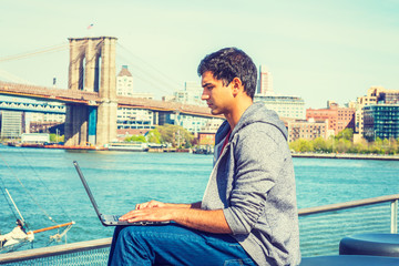 Fototapeta na wymiar Way to Success. East Indian American student travels, studies in New York, wearing hooded sweatshirt, jeans, sits by river, works on laptop computer, seriously thinks. Brooklyn bridge on background..