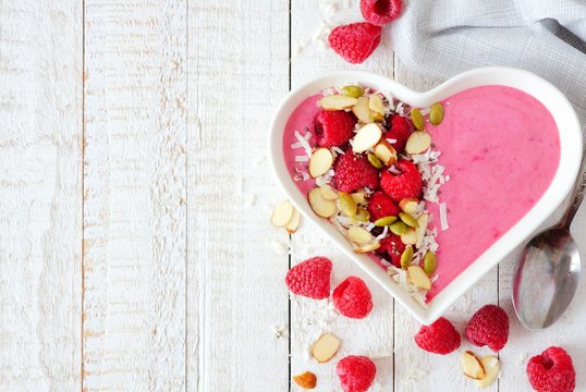 Healthy raspberry smoothie in a heart shaped bowl with almonds, pumpkin and chia seeds, coconut on a white wood background
