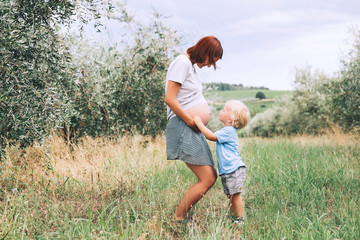 Child boy kissing belly of pregnant her mother on nature background.