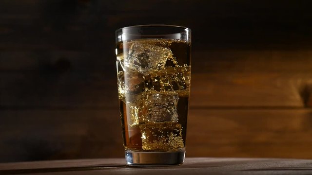 Glass of lemonade with ice cubes and bubbles. On dark wooden background. Rotating platform.