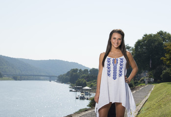 Beautiful young brunette woman standing in white dress along path near river with large suitcase