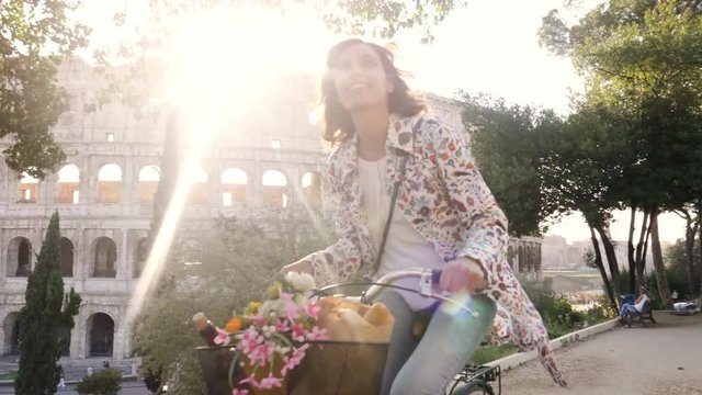 Beautiful young woman in colorful fashion riding bike in front of colosseum in Rome at sunset with trees happy attractive girl tourist with straw hat in colle oppio front view steadycam dolly