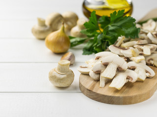 Fresh sliced mushrooms on chopping Board and onion and oil and parsley on white wooden table.