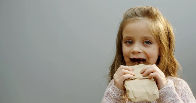 Portrait shot of the cute curly little girl standing in front of the camera and biting a chocolate bar with nuts on the white wall background. Close up. Inside