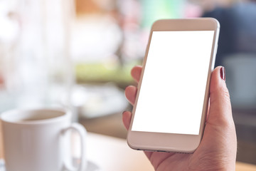 Mockup image of a woman's hand holding white mobile phone with blank desktop screen and coffee cup in cafe