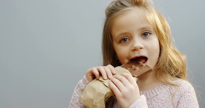 Close up of the blonde pretty small girl biting and eating a chocolate with nuts in front of the camera. White wall background. Indoor