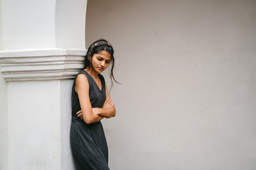 Attractive, fashionable and young Indian woman leaning against the wall of a white, colonial building and smiling in the day. 