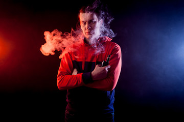 Portrait of a young handsome man holding a  vape and exhaling a large cloud of smoke on a dark background