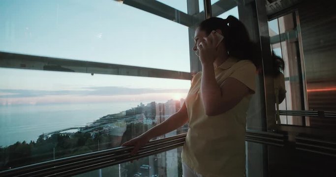 girl in the elevator with a panoramic view emotionally talking using a smartphone