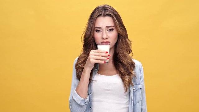 Displeased brunette woman in denim shirt refuses to drink kefir over yellow background
