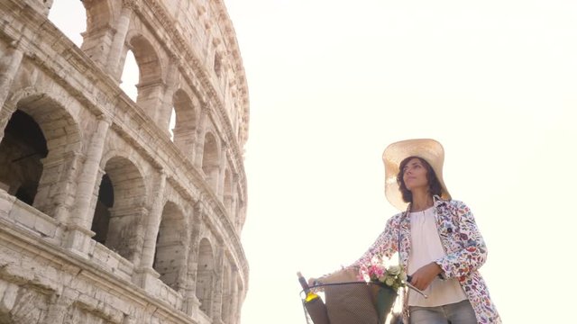 Beautiful young woman in colorful fashion dress walking alone with bike arriving in front of colosseum in Rome at sunset with happy attractive tourist girl with straw hat ground shot