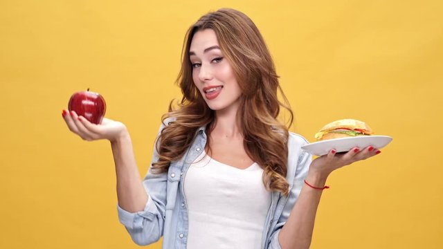 Sly Pretty brunette woman in denim shirt choosing between apple and burger and chooses an apple at the end over yellow background
