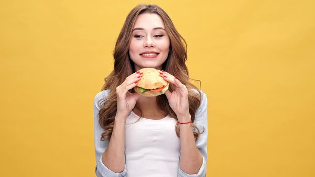 Pleased brunette woman in denim shirt eating big burger over yellow background
