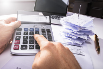 Person's Hand Calculating Invoice Near Bills And Laptop