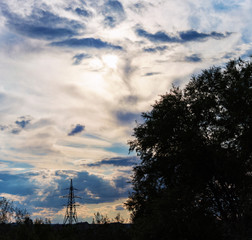 silhouettes of trees and a high-voltage tower against the background of dramatic clouds, through which the rays of the sun make their way