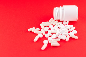 Close up white pill bottle with spilled out pills and capsules on scarlet red background with copy space. Focus on foreground, soft bokeh. Pharmacy drugstore concept