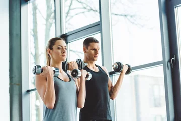 Papier Peint photo Lavable Fitness focused young man and woman exercising with dumbbells in gym
