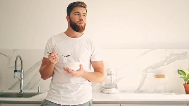 Pleased bearded man standing on kitchen and eating fruit while looking away
