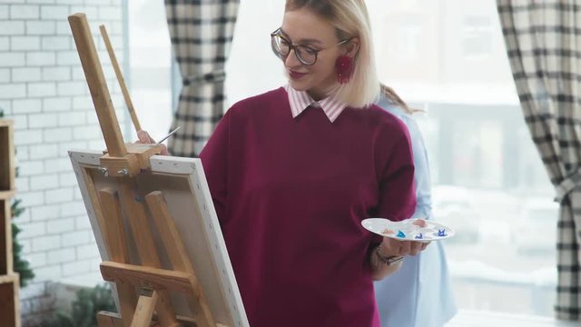 Two young beautiful women in stylish clothes are engaged in creativity and draw in a bright studio, standing at the easel. Two friends spend time together, laughing and painting.
