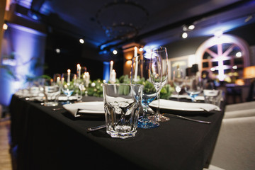 Glasses, forks, knives, napkins, candles and decorative flower on dark black tablecloth on table served for dinner in cozy restaurant. Empty served restaurant table with plates and wine glass.
