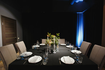 Glasses, forks, knives, napkins, candles and decorative flower on dark black tablecloth on table served for dinner in cozy restaurant. Empty served restaurant table with plates and wine glass.