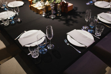 Close up glasses, forks, knives, napkins, candles and decorative flower on dark black tablecloth on table served for dinner in cozy restaurant. Empty served restaurant table with plates and wine glass