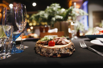 Sliced homemade meat products, cured meat, bacon, with fresh tomatoes, cucumbers on piece of wood, wine glasses on table with black tablecloth served for dinner in cozy luxury restaurant, close up.