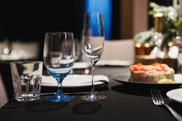 Close up three glasses, forks, knives, napkins, candles and decorative flower on dark black tablecloth on table served for dinner in cozy restaurant. Empty served restaurant table with wine glass.