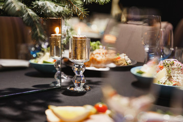 Burning candles, forks, knives, napkins, Glasses and decorative flower on dark black tablecloth on table served for dinner in cozy restaurant. Empty served restaurant table with plates and wine glass.