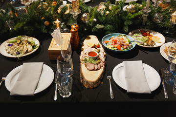 Glasses, forks, knives, napkins, candles and decorative flower on dark black tablecloth on table served for dinner in cozy restaurant. Food served restaurant table with plates and wine glass.